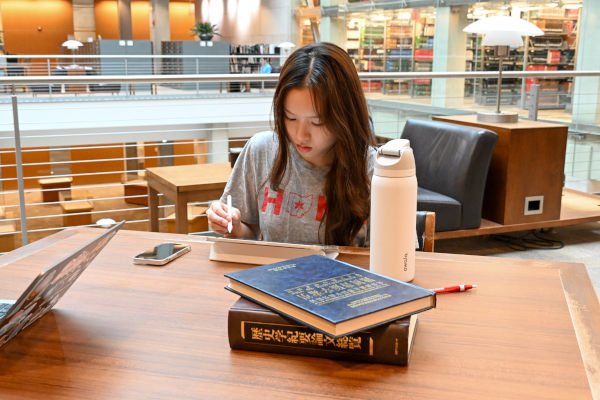 Person studying at a library table with books, laptop, and water bottle, surrounded by shelves and seating.